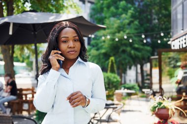 Telefon kullanarak sohbet ediyorlar. Beyaz gömlekli genç Afro-Amerikalı kadın yeşil ağaçların yanında ve iş binasının karşısında..