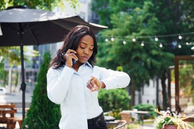 Telefon kullanarak sohbet ediyorlar. Beyaz gömlekli genç Afro-Amerikalı kadın yeşil ağaçların yanında ve iş binasının karşısında..
