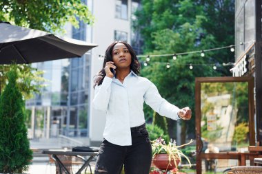 Telefon kullanarak sohbet ediyorlar. Beyaz gömlekli genç Afro-Amerikalı kadın yeşil ağaçların yanında ve iş binasının karşısında..