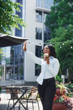 Bir bardak içkiyi el ele tutuşarak. Beyaz gömlekli genç Afro-Amerikalı kadın yeşil ağaçların yanında ve iş binasının karşısında..