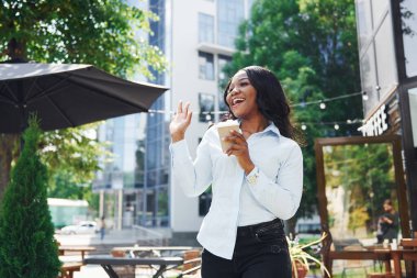 Bir bardak içkiyi el ele tutuşarak. Beyaz gömlekli genç Afro-Amerikalı kadın yeşil ağaçların yanında ve iş binasının karşısında..
