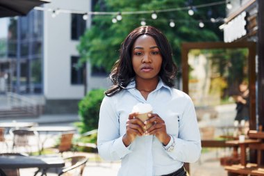 Bir bardak içkiyi el ele tutuşarak. Beyaz gömlekli genç Afro-Amerikalı kadın yeşil ağaçların yanında ve iş binasının karşısında..