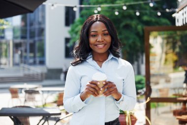 Bir bardak içkiyi el ele tutuşarak. Beyaz gömlekli genç Afro-Amerikalı kadın yeşil ağaçların yanında ve iş binasının karşısında..