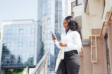 Ellerinde telefonla. Beyaz gömlekli genç Afro-Amerikan kadın şehir dışında iş binasına karşı..