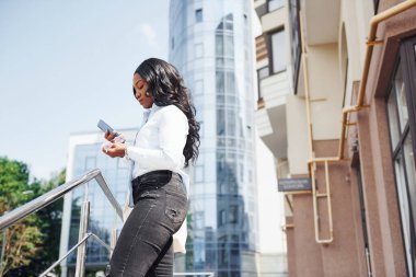 Ellerinde telefonla. Beyaz gömlekli genç Afro-Amerikan kadın şehir dışında iş binasına karşı..