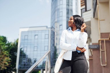 Ellerinde telefonla. Beyaz gömlekli genç Afro-Amerikan kadın şehir dışında iş binasına karşı..