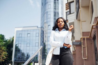 Ellerinde telefonla. Beyaz gömlekli genç Afro-Amerikan kadın şehir dışında iş binasına karşı..