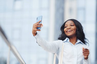 Ellerinde telefonla. Beyaz gömlekli genç Afro-Amerikan kadın şehir dışında iş binasına karşı..