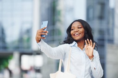 Ellerinde telefonla. Beyaz gömlekli genç Afro-Amerikan kadın şehir dışında iş binasına karşı..