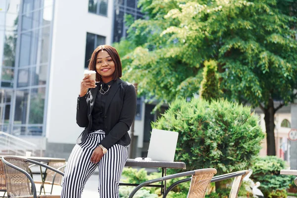 Laptop ve masa. Şehirde, yeşil ağaçların yanında ve iş inşaatına karşı şık giyinmiş Afro-Amerikalı genç bir kadın..