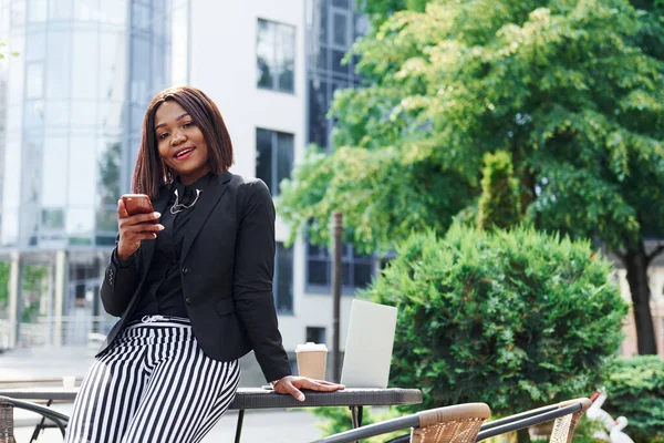 Laptop ve masa. Şehirde, yeşil ağaçların yanında ve iş inşaatına karşı şık giyinmiş Afro-Amerikalı genç bir kadın..