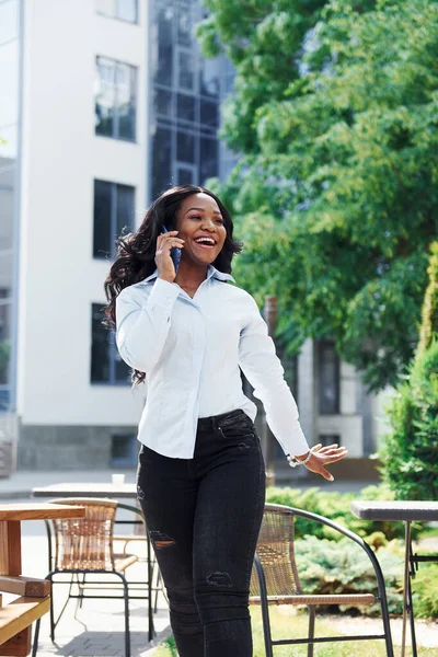 Telefon kullanarak sohbet ediyorlar. Beyaz gömlekli genç Afro-Amerikalı kadın yeşil ağaçların yanında ve iş binasının karşısında..