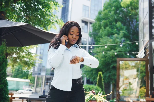 Telefon kullanarak sohbet ediyorlar. Beyaz gömlekli genç Afro-Amerikalı kadın yeşil ağaçların yanında ve iş binasının karşısında..