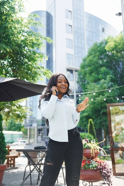 Telefon kullanarak sohbet ediyorlar. Beyaz gömlekli genç Afro-Amerikalı kadın yeşil ağaçların yanında ve iş binasının karşısında..
