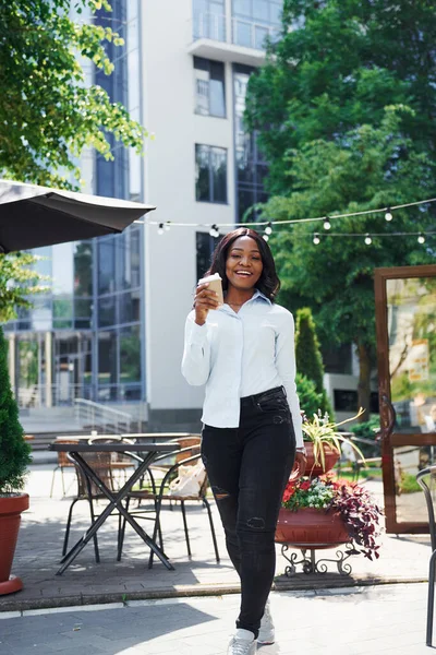 Bir bardak içkiyi el ele tutuşarak. Beyaz gömlekli genç Afro-Amerikalı kadın yeşil ağaçların yanında ve iş binasının karşısında..