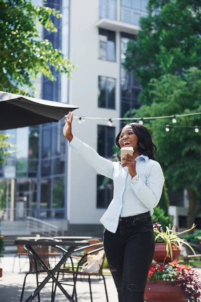Bir bardak içkiyi el ele tutuşarak. Beyaz gömlekli genç Afro-Amerikalı kadın yeşil ağaçların yanında ve iş binasının karşısında..