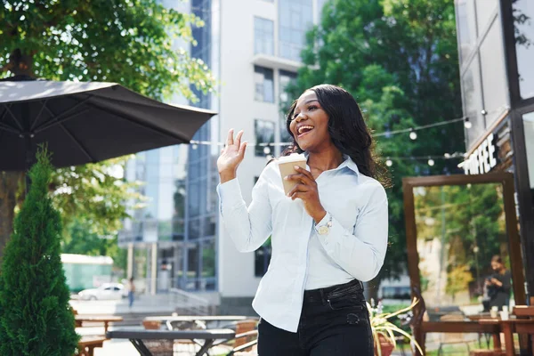 Bir bardak içkiyi el ele tutuşarak. Beyaz gömlekli genç Afro-Amerikalı kadın yeşil ağaçların yanında ve iş binasının karşısında..