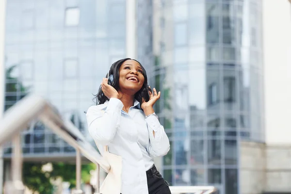 用耳机听音乐 年轻的非洲裔美国女人穿着白衬衫站在城中的商业楼前 — 图库照片