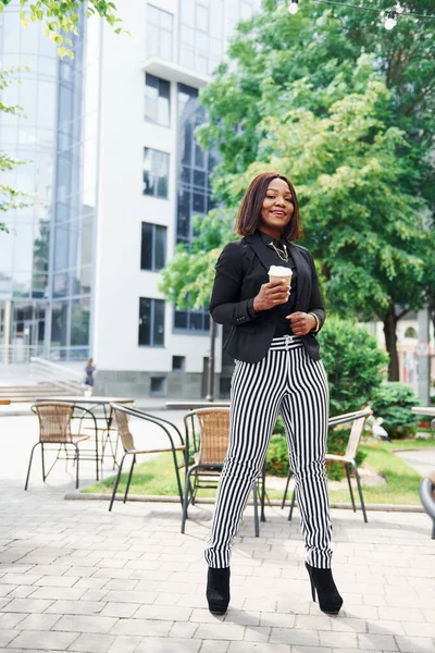 Kahve molası. Şehirde, yeşil ağaçların yanında ve iş inşaatına karşı şık giyinmiş Afro-Amerikalı genç bir kadın..