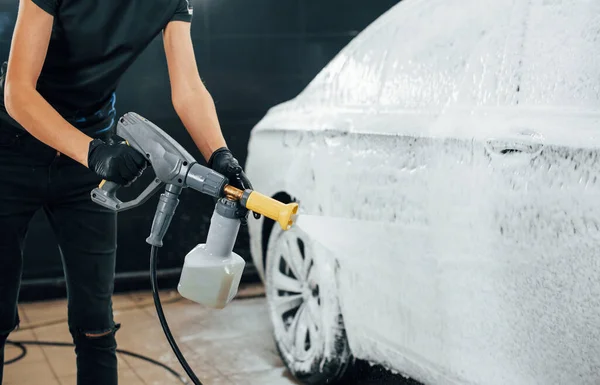 Using high pressure water. Modern black automobile get cleaned by woman inside of car wash station.