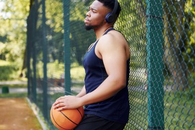 Kablosuz kulaklıklı Afrikalı Amerikalı adam mola veriyor ve sahada topla metal ağlara yaslanıyor..