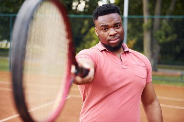 Pembe gömlekli Afro-Amerikan bir adam dışarıda tenis raketiyle kameraya poz veriyor..