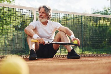 Yerde oturuyor ve mola veriyor. Gündüz vakti tenis kortunda raketi olan son sınıf modern stil sahibi bir adam..