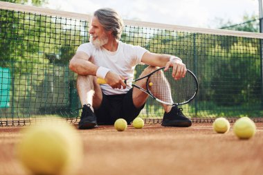 Yerde oturuyor ve mola veriyor. Gündüz vakti tenis kortunda raketi olan son sınıf modern stil sahibi bir adam..