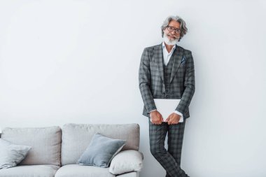 Kıdemli stil sahibi, gri saçlı, sakallı, elinde dizüstü bilgisayarla duvarın yanında duran modern bir adam..