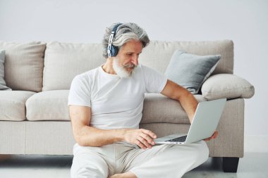 Dizüstü bilgisayarla kanepede oturur ve müzik dinler. Gri saçlı ve sakallı son sınıf modacı modern adam..