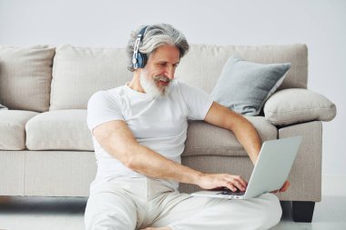 Dizüstü bilgisayarla kanepede oturur ve müzik dinler. Gri saçlı ve sakallı son sınıf modacı modern adam..