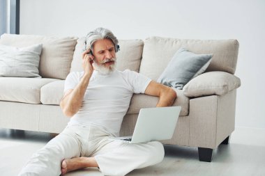 Dizüstü bilgisayarla kanepede oturur ve müzik dinler. Gri saçlı ve sakallı son sınıf modacı modern adam..