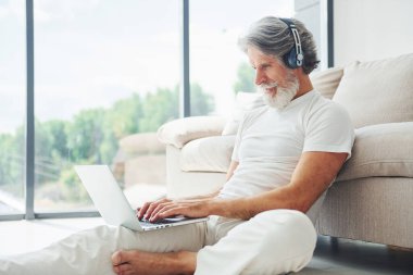 Dizüstü bilgisayarla kanepede oturur ve müzik dinler. Gri saçlı ve sakallı son sınıf modacı modern adam..