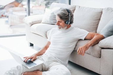 Dizüstü bilgisayarla kanepede oturur ve müzik dinler. Gri saçlı ve sakallı son sınıf modacı modern adam..