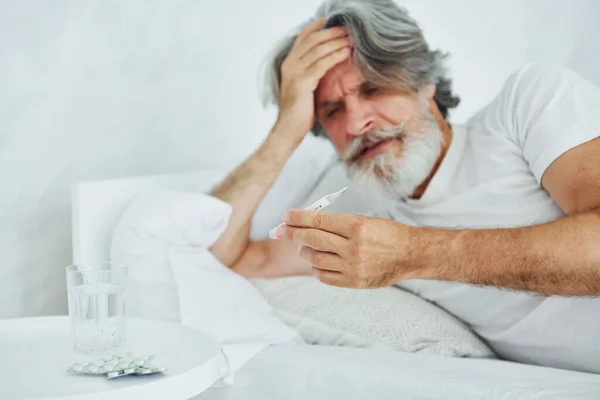 Feels sick. On the bed. Senior stylish modern man with grey hair and beard indoors.