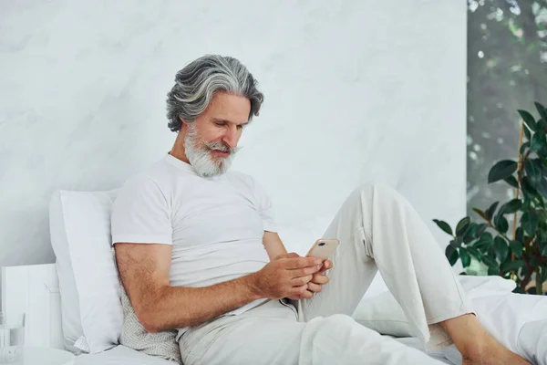 Sits on bed and using phone. Senior stylish modern man with grey hair and beard indoors.