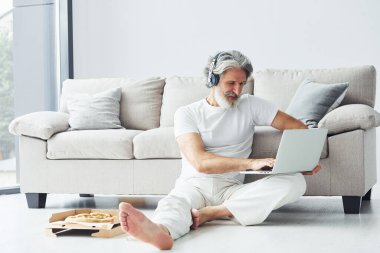 Kulaklıkla. Kanepenin yanında oturuyor. Gri saçlı ve sakallı son sınıf modacı modern adam..