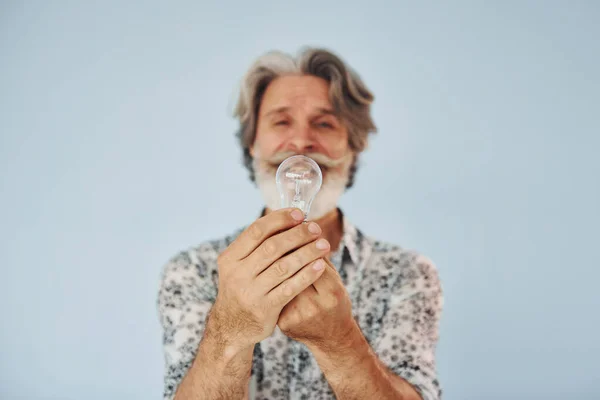 Holds light bulb in hands. Conception of ideas and inspiration. Senior stylish modern man with grey hair and beard indoors.