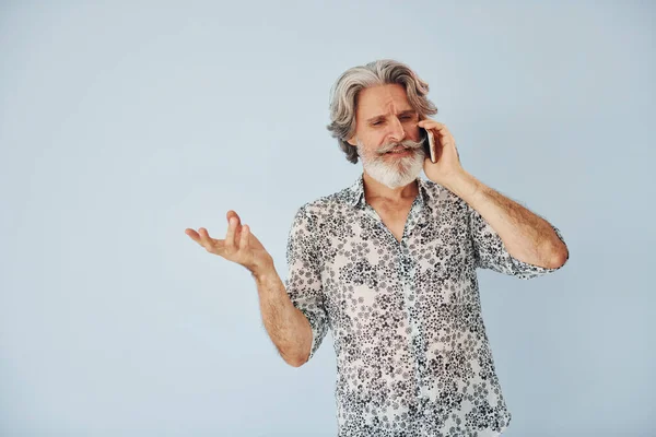 Having call when standing against wall. Senior stylish modern man with grey hair and beard indoors.