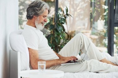 Yatakta dizüstü bilgisayarlı serbest çalışan. Gri saçlı ve sakallı son sınıf modacı modern adam..