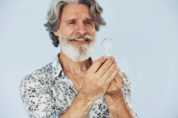 Holds light bulb in hands. Conception of ideas and inspiration. Senior stylish modern man with grey hair and beard indoors.
