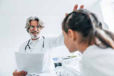 Küçük kıza yapılan muayenenin sonuçlarını okuyorum. Beyaz önlüklü, gri saçlı ve sakallı kıdemli erkek doktor klinikte..