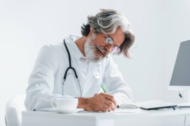 Bilgisayar masasında yazıyor. Beyaz önlüklü, gri saçlı ve sakallı kıdemli erkek doktor klinikte..
