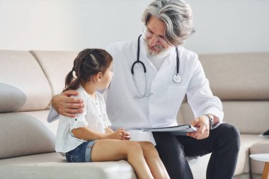 Küçük kızla konuşuyor. Müşteriyle çalışıyorum. Beyaz önlüklü, gri saçlı ve sakallı kıdemli erkek doktor klinikte..