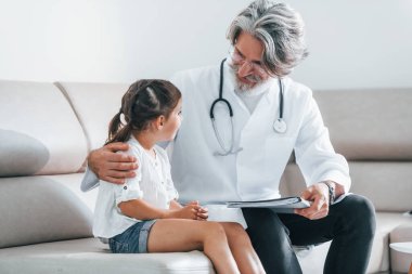 Küçük kızla konuşuyor. Müşteriyle çalışıyorum. Beyaz önlüklü, gri saçlı ve sakallı kıdemli erkek doktor klinikte..