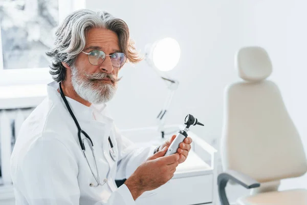 Portrait of senior male doctor with grey hair and beard in white coat is indoors in otorhinolaryngology clinic.