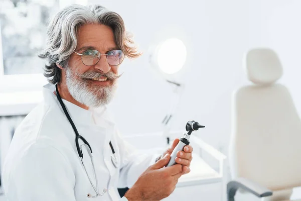 Portrait of senior male doctor with grey hair and beard in white coat is indoors in otorhinolaryngology clinic.
