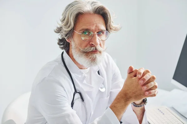 Portrait of senior male doctor with grey hair and beard in white coat is indoors in clinic.