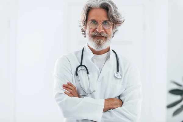 Portrait of senior male doctor with grey hair and beard in white coat standing indoors in clinic.