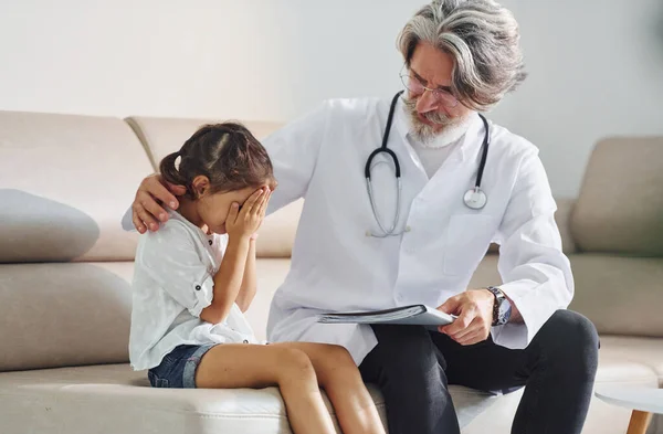 Talks with little sad girl. Working with customer. Senior male doctor with grey hair and beard in white coat is indoors in clinic.
